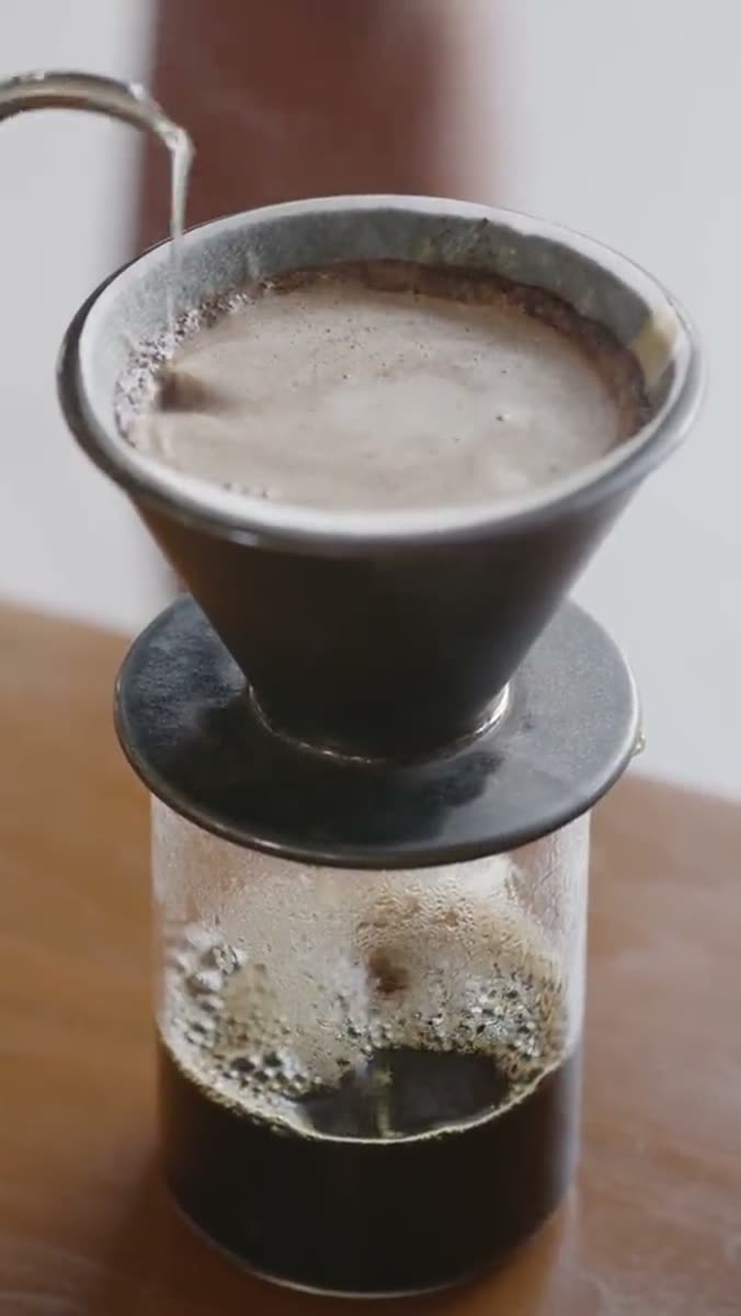 A gooseneck kettle pouring a thin stream of water into a dark ceramic pour-over cone full of coffee bloom, sitting on a glass carafe half full of coffee, on a wooden table