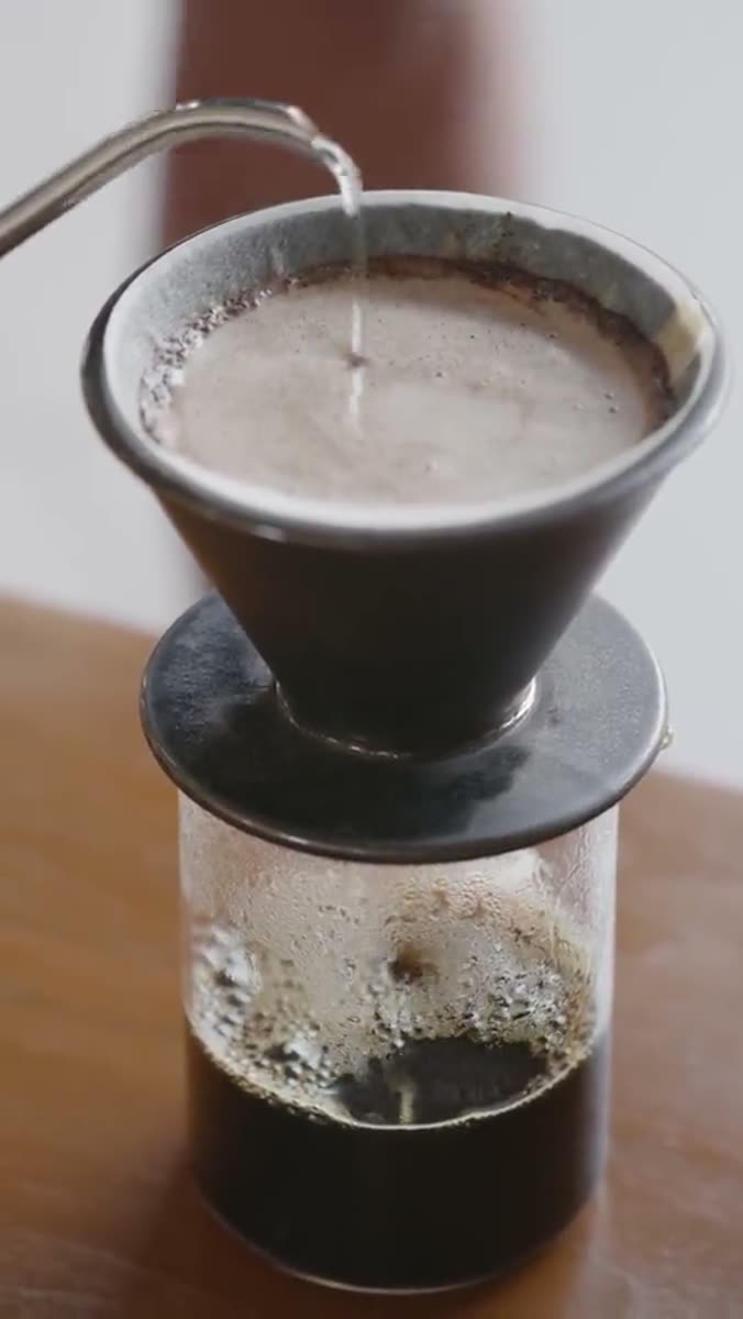 Water poured from a gooseneck kettle into a ceramic dripper sitting on a glass carafe, on a wooden table in daylight