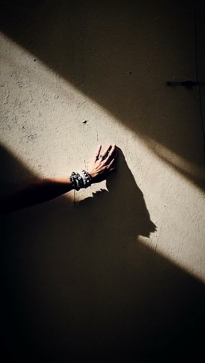 A hand and forearm wearing beaded bracelets pressed flat against a sunlit plaster wall, with a hard diagonal shadow crossing the frame