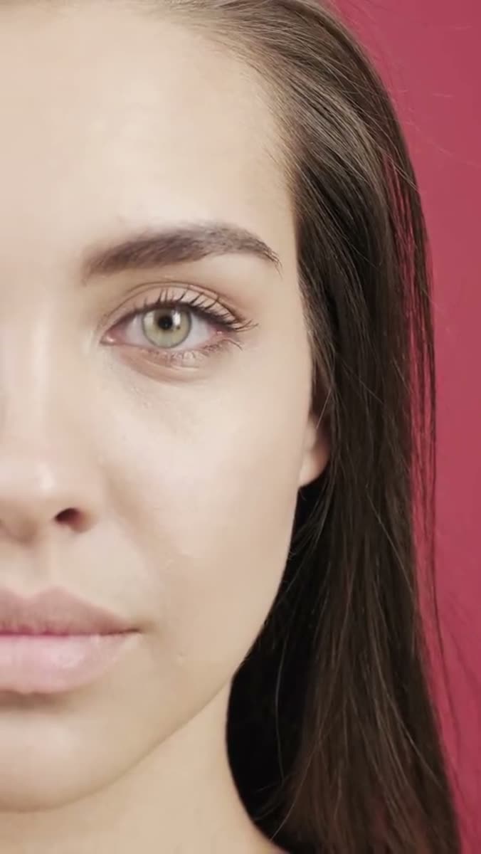 Half a woman's face in close crop against a flat deep pink background, one green eye and a bare lip visible