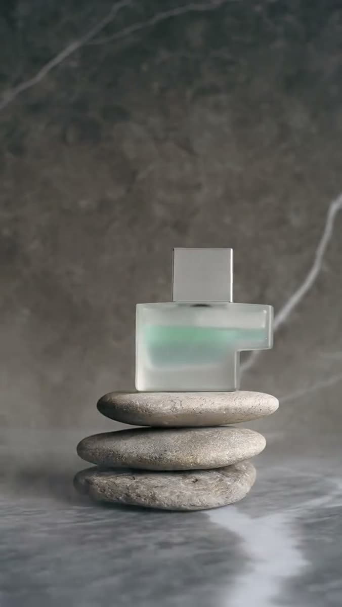A frosted glass perfume bottle with a mirror-polished silver cap balanced on three stacked river stones against a gray marble wall