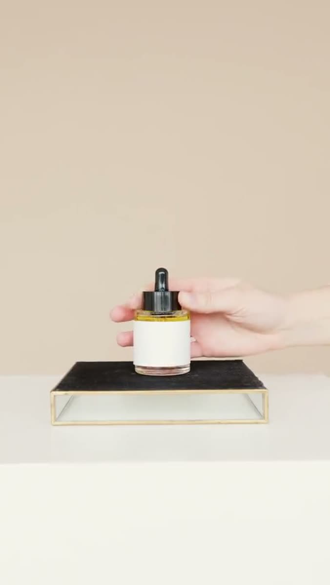 A hand lifting a small glass dropper bottle with a black cap off a dark stone tray on a pale table, against a beige wall