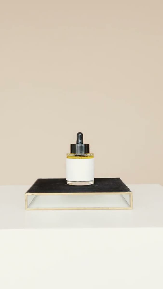 A glass dropper bottle with a white label standing on a dark slate tile inside a brass-edged tray on a pale plinth