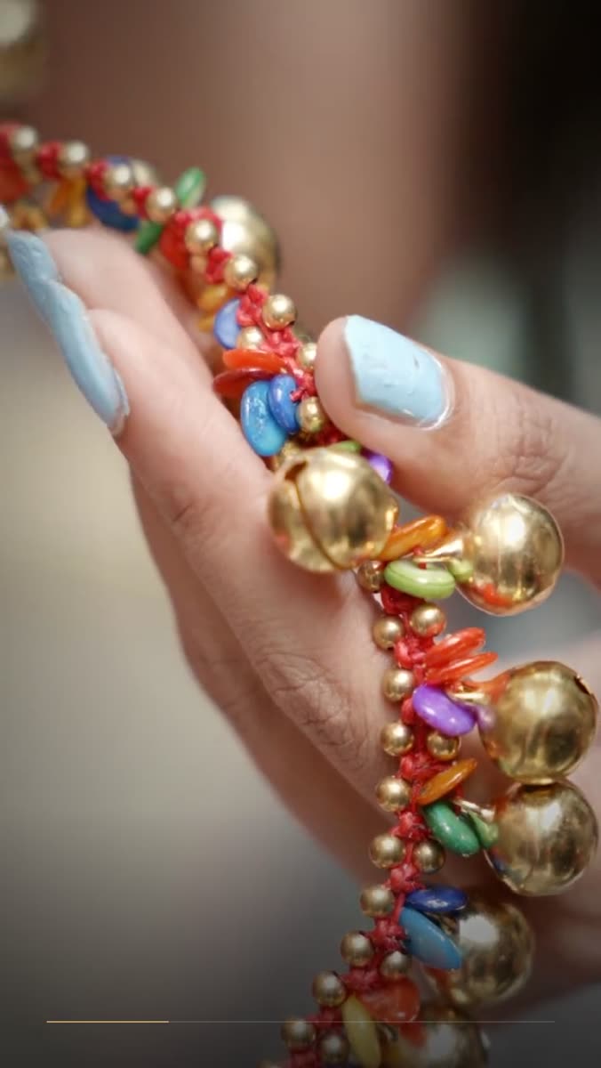 A hand with pale blue nail polish holding a beaded necklace made of polished gold spheres and small colored glass beads strung on a red cord, in soft daylight