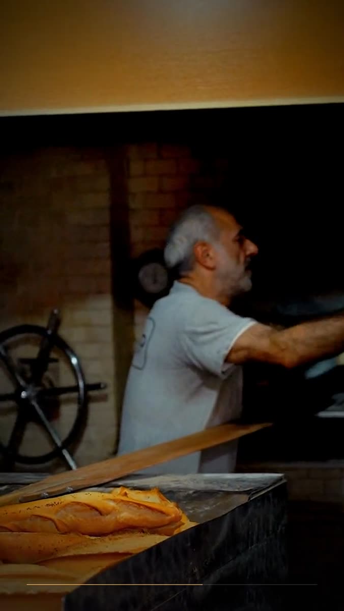 A baker in a white shirt turning away from a brick oven, a wooden peel angled across the foreground with raw dough on it, warm tungsten light on the ceiling above