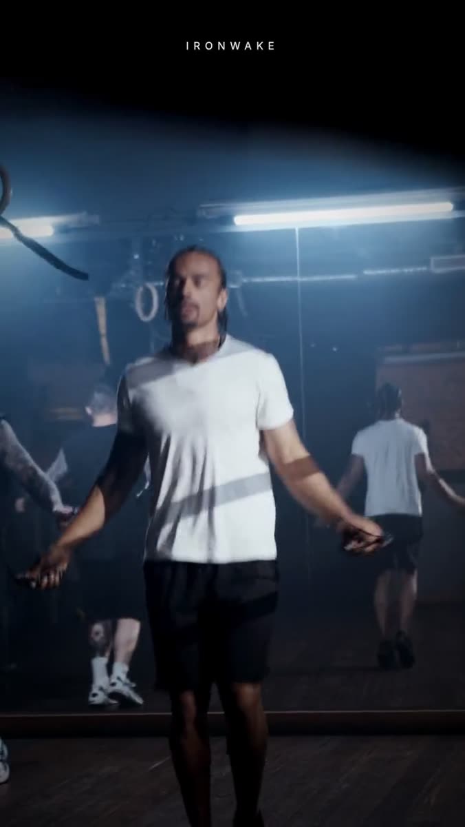 A man in a white t-shirt skipping rope in a dark gym filled with haze, other people skipping behind him in soft focus, a small letterspaced wordmark set across the very top of the frame