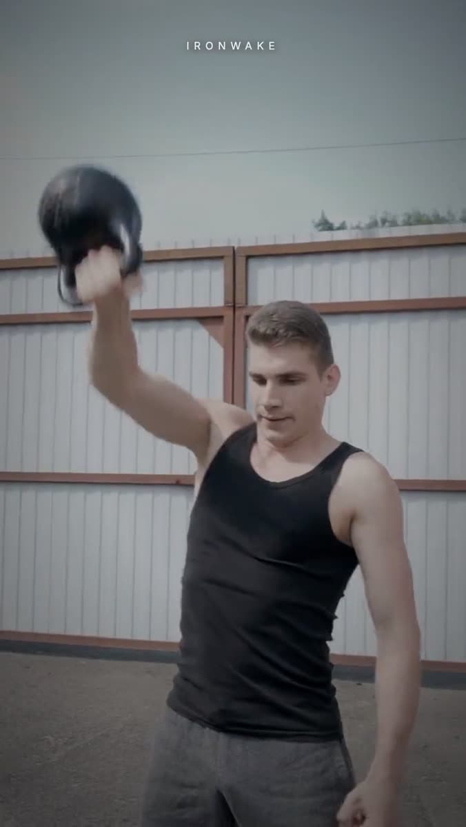 A man in a black vest swinging a black kettlebell overhead outdoors in front of a white corrugated wall