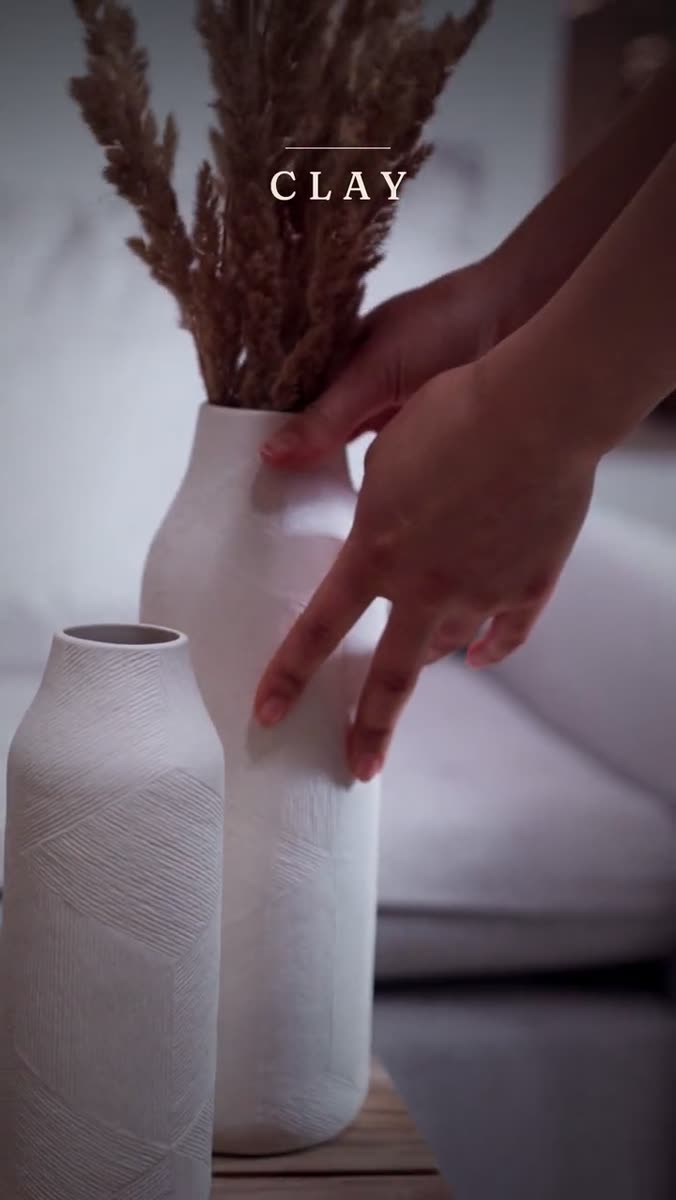 A hand steadying a tall white ceramic vase of dried pampas grass beside a second ribbed vase, with the word CLAY set above them
