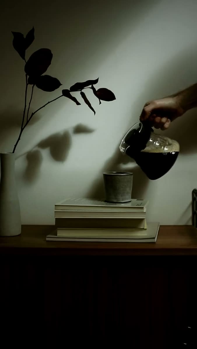A hand tips a glass carafe of dark coffee toward a stoneware cup on a stack of books, lit hard from one side, with the shadow of a bare branch across the wall behind