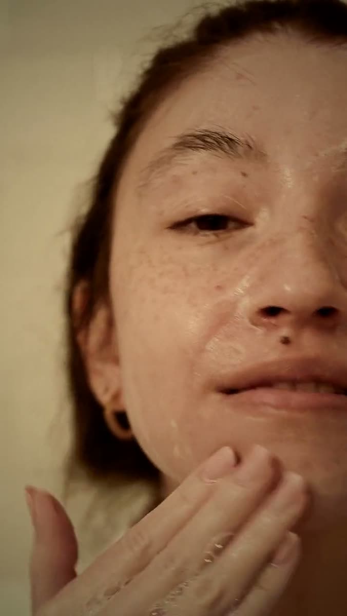 Close crop of a woman's face at a bathroom mirror, water and cleanser on her cheek, one hand rising into the bottom of the frame