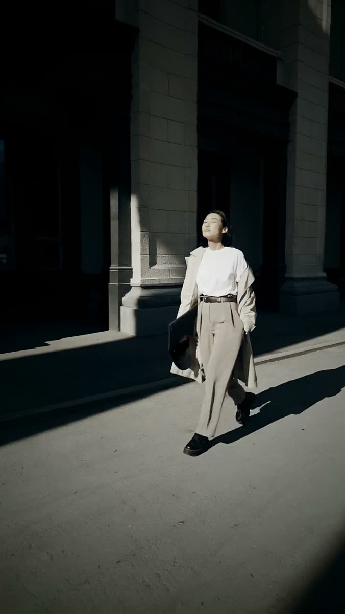 A woman in a white top, tan wide-leg trousers and an open coat walking through hard low sun past a colonnade, casting a long shadow across the pavement