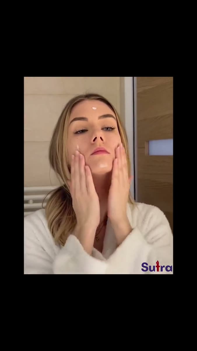 A woman in a white robe pressing both palms to her cheeks in a tiled bathroom, shot square to camera