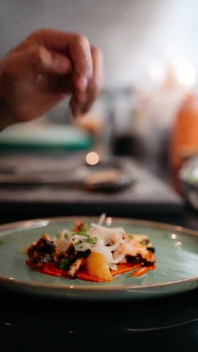 A hand held above a sage green ceramic plate with fingers pinched, releasing shredded garnish over grilled fish, shaved onion, red sauce and a wedge of orange, in a dark dining room with a candle out of focus behind