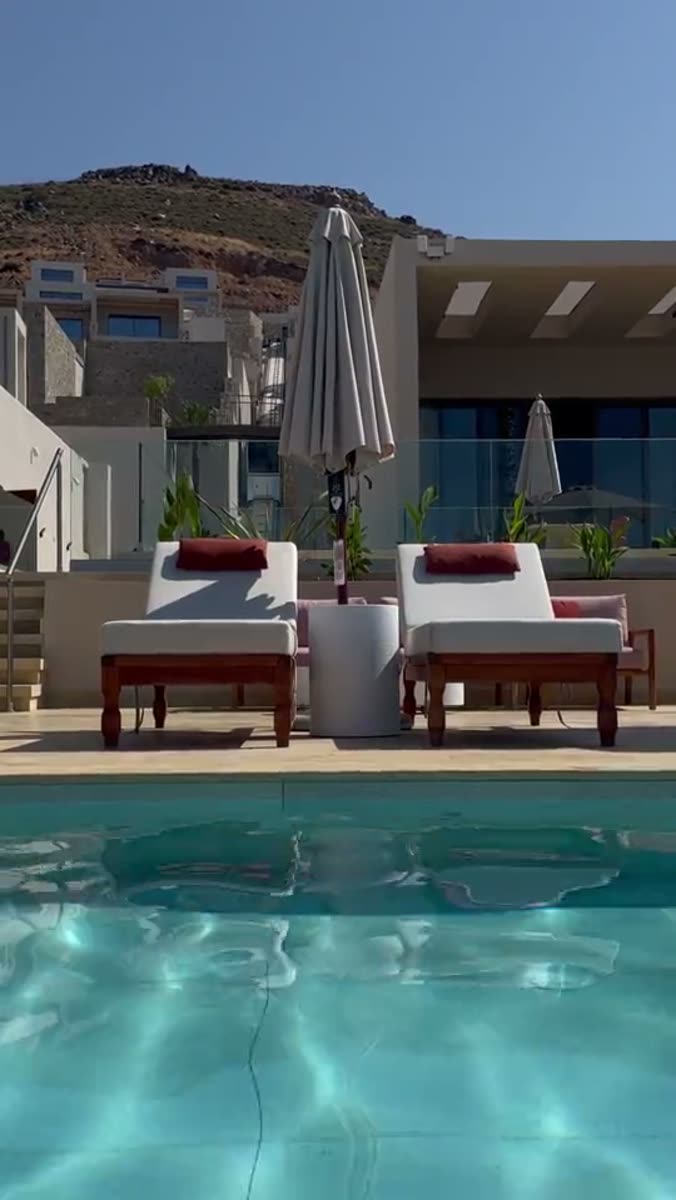 Two wooden sun loungers with white cushions and dark red bolsters flanking a closed cream parasol, seen across a turquoise pool, with white terraced villas on a dry hillside behind