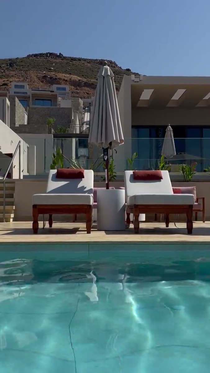 A hotel pool terrace in bright sun: two wooden sun loungers with white cushions and red bolsters, a closed cream parasol between them, a glass balustrade and a stepped white building behind, and a dry rocky hillside above