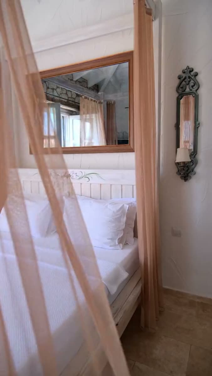 A canopied bed seen through hanging sheer drapes, a wood-framed mirror above the headboard reflecting a curtained window and a beamed ceiling, an iron candle sconce on a plaster wall, stone floor