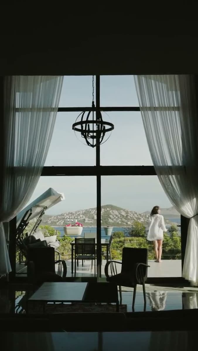 A dark interior looking out through a tall black window frame to a terrace, sheer curtains at both sides and a small figure in white at the railing above a bay